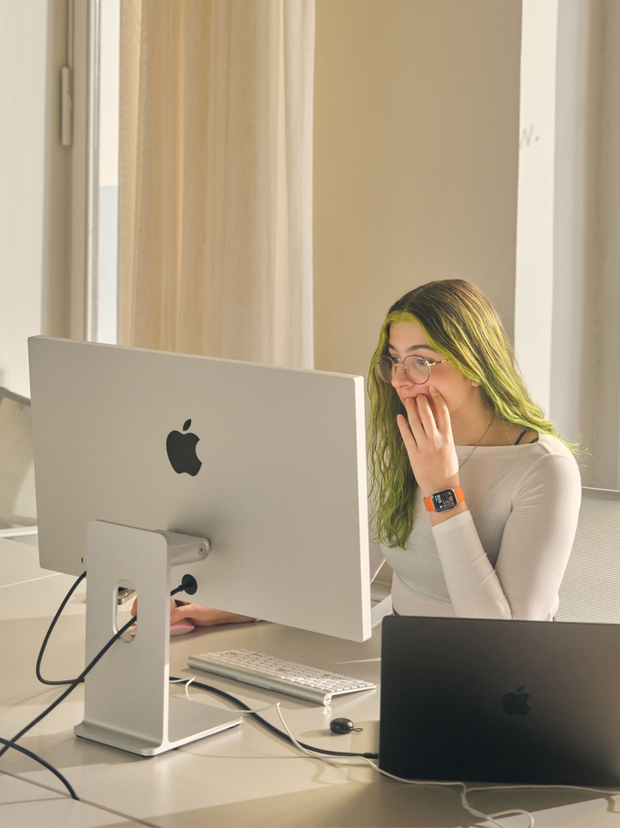 Person with green hair working at a desk with an Apple computer and laptop in a sunlit office
