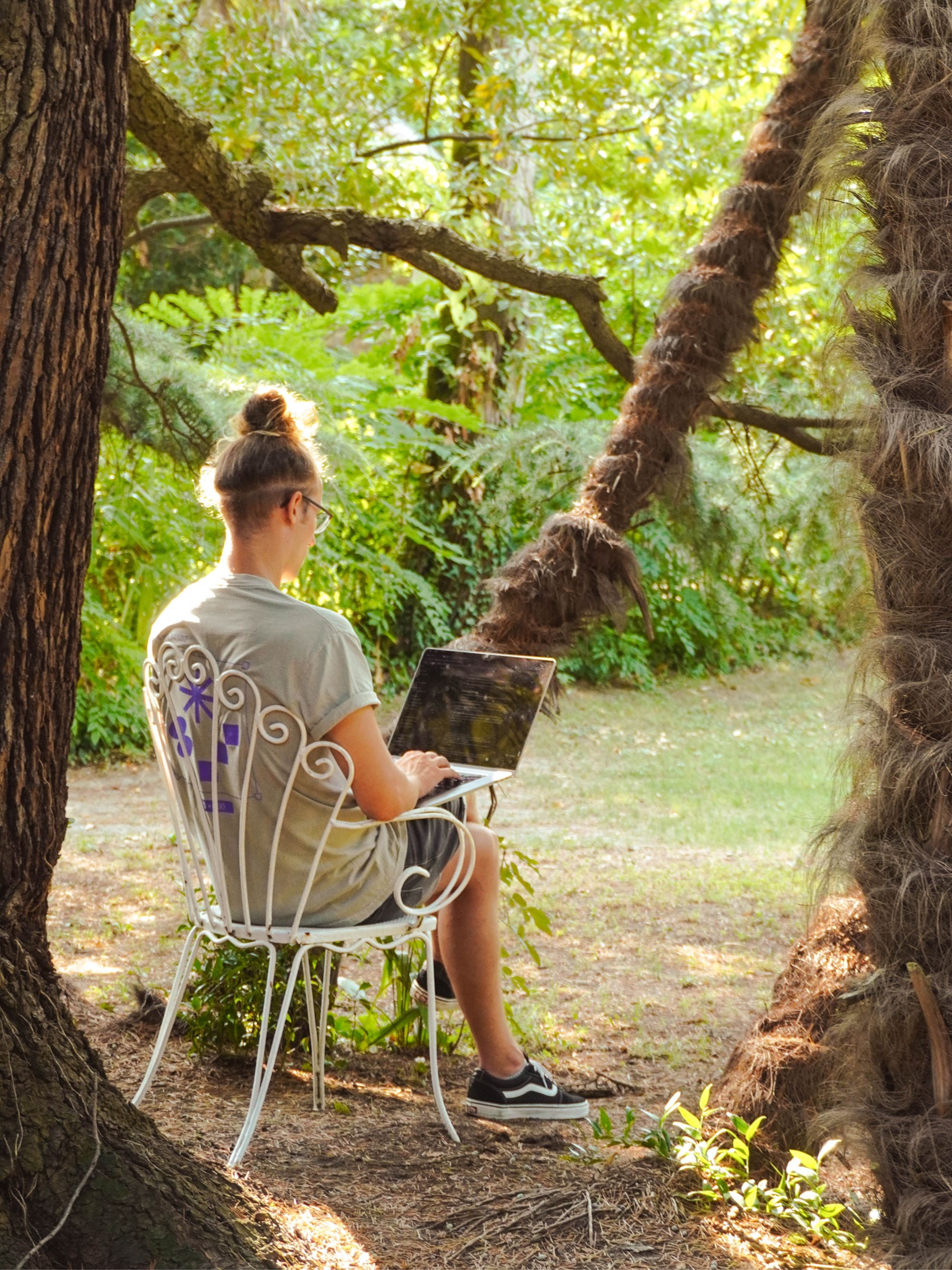 Person sitting on a white metal chair outdoors, working on a laptop surrounded by greenery