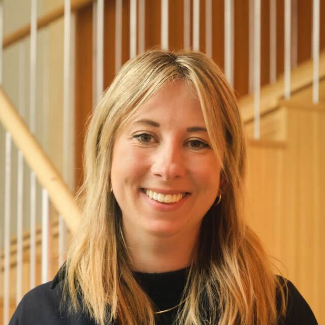 Person with shoulder-length blonde hair, wearing a black top, standing in front of a wooden staircase
