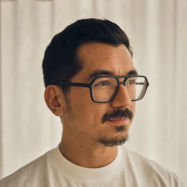 Person with short dark hair and glasses, wearing a white shirt, standing in front of light curtains
