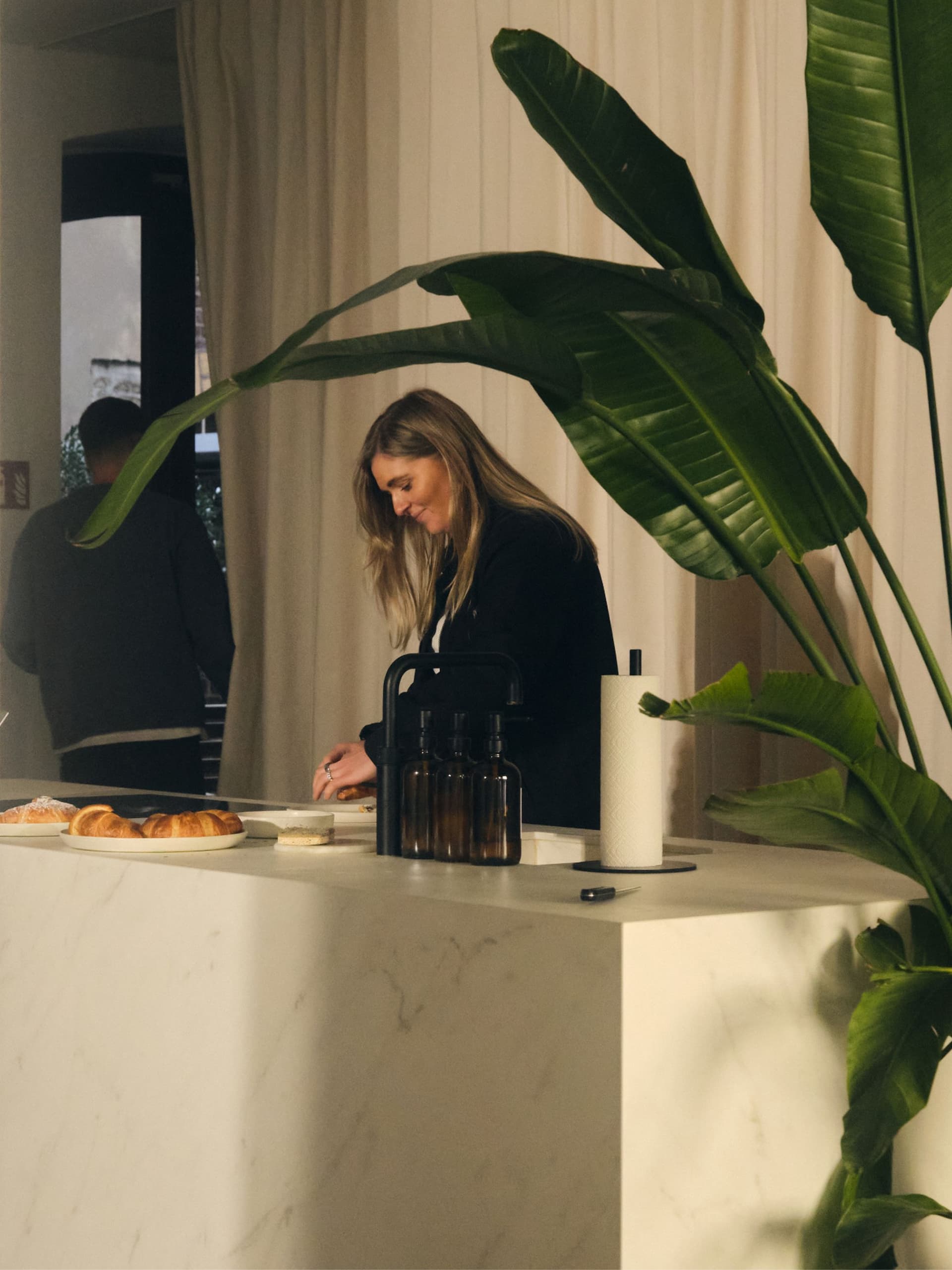 Person preparing food at a kitchen counter with pastries and bottles, next to a large plant