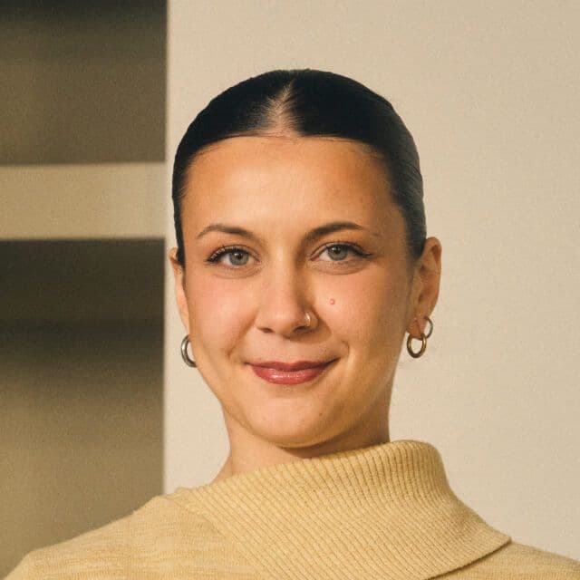 Person with dark hair tied back, wearing a beige turtleneck sweater and hoop earrings, standing indoors