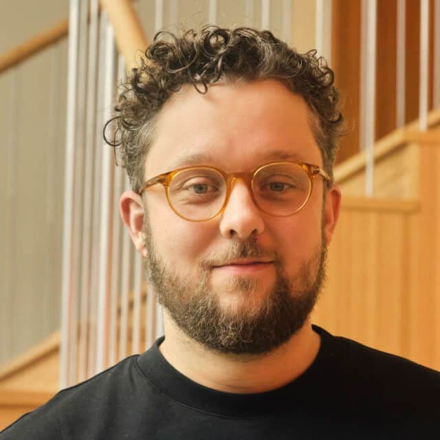 Person with short curly hair and beard, wearing a black shirt, standing in front of a wooden staircase